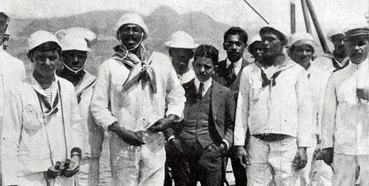 O líder da Revolta da Chibata, João Cândido (à esquerda do homem com terno escuro), com repórteres, oficiais e marinheiros a bordo do Minas Gerais. Créditos: autoria desconhecida. joão cândido na revolta da chibata